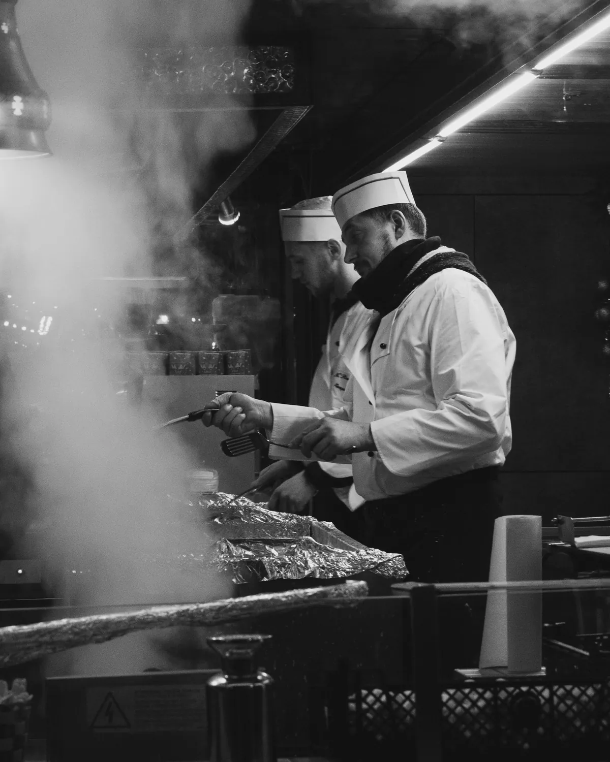 Två kockar i kockmössa lagar mat över öppen grill medan ånga stiger, i svartvitt